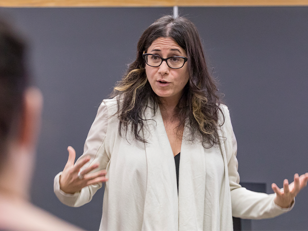 A woman instructor stands in front of a classroom, gesticulating with her hands and speaking to the students.